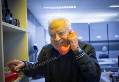 O jornalista Cid Moreira, então com 89 anos, posa sorrindo segurando um telefone rente à orelha